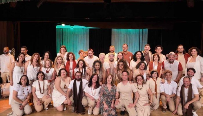 grupo com os integrantes do Coral Municipal de Jundiaí reunidos no palco, vestidos em tons claros e sorrindo, posados em várias fileiras diante de um fundo i