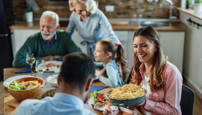 Young happy woman laughing while passing food to her husband and enjoying in family lunch at dining table.